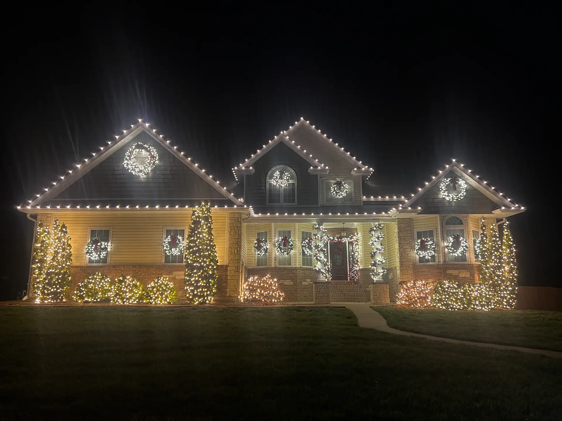 A two-story home glowing with warm white rooflines, lit wreaths in the windows, and wrapped trees along the front